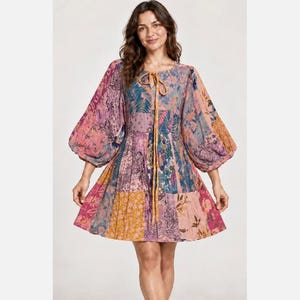 May include: A short-length dress featuring a patchwork design with various floral and paisley patterns in shades of pink, purple, orange, and blue. The dress has long, voluminous puff sleeves, a round neckline with a tie closure, and a flared skirt.