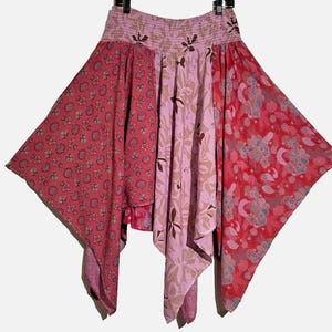 May include: A handkerchief-hem skirt featuring a smocked elastic waistband and a patchwork design. The garment displays various shades of pink and red with floral and abstract patterns, creating a layered, asymmetrical silhouette with pointed edges.