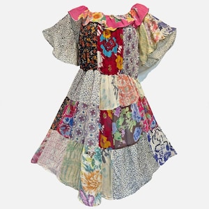 May include: A patchwork dress with a ruffled neckline and short, flared sleeves. The dress features a variety of fabric patterns and colors, including floral, paisley, and geometric designs. The dress has a tiered skirt and a pink neckline.