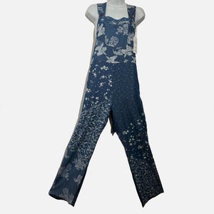 May include: Blue denim overalls featuring a patchwork design with various white floral and botanical patterns. The garment includes a front bib pocket, wide shoulder straps, and long pant legs with a consistent blue and white color scheme throughout.