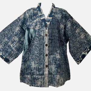 May include: A blue and white patterned jacket with a V-neckline and five brown buttons down the front. The garment features a mix of abstract textures, including vertical lines and circular motifs on the wide sleeves, with a structured collar.