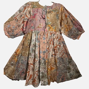 May include: A knee-length dress featuring a patchwork design of various floral and paisley patterns in muted tones of peach, brown, tan, and soft blue. The garment has long, voluminous puff sleeves, a round neckline with a tie closure, and a gathered waist.