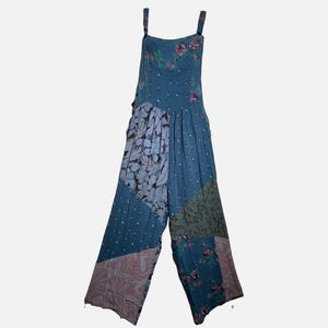 May include: A blue patchwork jumpsuit featuring a mix of floral, polka dot, and leaf patterns in shades of pink, green, and white. The garment has a square neckline, thin shoulder straps, and wide legs with a varied panel design throughout.