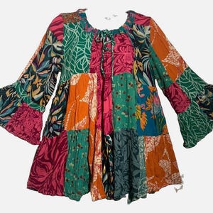 May include: A long-sleeved tunic featuring a patchwork design with various floral and leaf patterns in vibrant shades of orange, hot pink, teal, and dark green. The garment has a round neckline with a drawstring tie closure and flared bell sleeves.