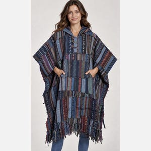 May include: A hooded poncho featuring a patchwork design with vertical stripes and geometric patterns in shades of blue, brown, and maroon. The garment includes a front kangaroo pocket, wooden toggle closures at the neckline, and a fringed hemline.