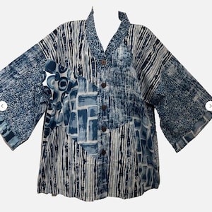 May include: A button-down shirt featuring a patchwork design of various blue and white patterns, including vertical stripes, abstract brushstrokes, and circular motifs. The garment has a V-neckline, wide three-quarter length sleeves, and brown buttons.