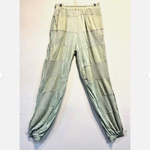 May include: Light green patchwork pants with an elastic waistband and gathered ankles. The pants are made of a soft, flowing fabric and are hanging on a hanger against a white wall. The pants have a relaxed fit.