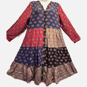 May include: A long-sleeved, button-down tiered dress featuring a patchwork design with various floral patterns in shades of red, blue, black, and muted tan. The garment has a V-neckline and a loose, flowing silhouette with multiple horizontal fabric panels.