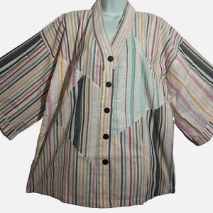 May include: A button-down shirt featuring a multi-directional striped pattern in shades of black, pink, yellow, and teal on a cream background. The garment has a V-neckline, wide three-quarter length sleeves, and five dark brown circular buttons.