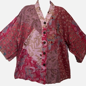 May include: A button-down shirt featuring a patchwork design in various shades of pink, red, and burgundy. The garment displays a mix of floral, paisley, and leaf patterns across its panels, with a V-neckline and five dark brown circular buttons.