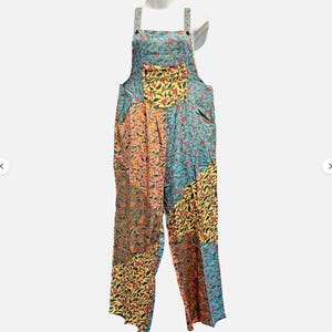 May include: A pair of full-length overalls featuring a patchwork design with various floral patterns in shades of blue, yellow, pink, and green. The garment includes a square chest pocket, two side pockets, and adjustable shoulder straps.