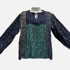 May include: A long-sleeved blouse featuring a patchwork design with various floral and geometric patterns in shades of navy blue, green, and cream. The garment has a round neckline with a small tie closure and gathered fabric at the shoulders.