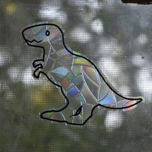 May include: A holographic sticker of a Tyrannosaurus Rex dinosaur in a walking pose. The sticker is outlined in black and has a rainbow iridescent finish.