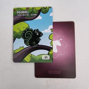 May include: A card featuring a black squirrel illustration perched on a tree branch, with the word "SQUIRREL" at the top. The background shows a blue sky and green foliage. A second card with a unicorn design is visible.
