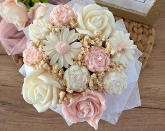 Puede incluir: Un ramo de velas de flores de cera blancas y rosas dispuestas en un envoltorio de papel blanco. Las velas tienen forma de rosas, peonías y margaritas.