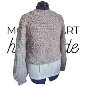 Peut inclure: Un pull crocheté fait main, rose clair et gris, avec un motif de point texturé. Le pull a des manches longues et une encolure ronde. Les mots "MO" et "art handmade" sont écrits en script noir sur les côtés gauche et droit de l'image.