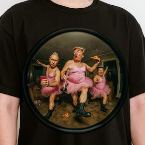 May include: Black t-shirt featuring a circular graphic of three figures in pink tutus and dresses, holding pizza and drinks. The graphic is framed in a dark circle. The word "BLACK" is printed at the bottom of the shirt.