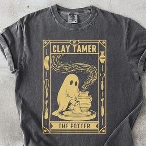 Könnte beinhalten: Dunkelgraues T-Shirt mit einem goldenen Aufdruck, der einen Geist zeigt, der Ton auf einer Töpferscheibe formt. Der Text lautet "CLAY TAMER" und "THE POTTER". Die Ärmel sind hochgekrempelt.