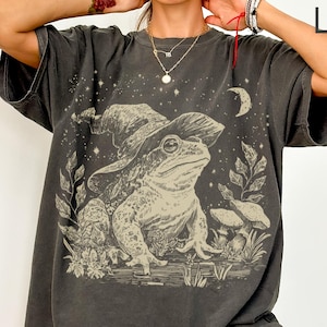 May include: Dark gray t-shirt featuring a whimsical illustration of a frog wearing a witch's hat, set against a night sky with a crescent moon and stars. The design includes plants and mushrooms, creating a magical, nature-inspired theme. The word "TEEZARO" is printed on the top right.