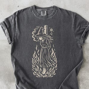 May include: Dark gray t-shirt featuring a witch illustration. The witch is depicted in a long dress and witch hat, surrounded by flames. The design is in a cream color. The word "PEPPER" is printed at the bottom of the shirt.