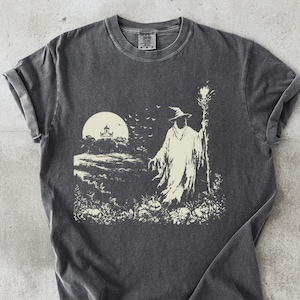 May include: Dark grey t-shirt with a white graphic of a wizard holding a staff, set against a full moon and castle backdrop. The shirt has a vintage, distressed appearance. The word "TEEZARO" is in the upper right corner.
