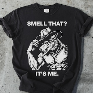 Puede incluir: Camiseta negra con un diseño blanco de un lobo musculoso con sombrero de vaquero. El texto "SMELL THAT? IT'S ME." está impreso encima y debajo de la ilustración. La camiseta está sobre una superficie gris.