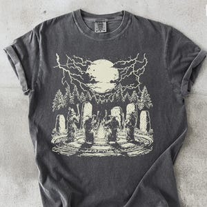 May include: Dark grey t-shirt featuring a cream-colored graphic of a full moon, lightning, trees, and figures in a circle. The shirt has rolled-up sleeves and a crew neck. The word "PEPPER" is at the bottom.