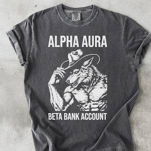 May include: Dark gray t-shirt with a white graphic of a muscular wolf wearing a cowboy hat. The text "ALPHA AURA" is above the wolf, and "BETA BANK ACCOUNT" is below. The shirt is made of a soft material.