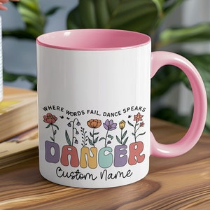 May include: White ceramic mug with a pink interior and handle. The mug features the text "Where words fail, dance speaks" and the word "Dancer" in colorful letters, with floral illustrations. The bottom of the mug says "Custom Name".