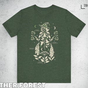 Puede incluir: Camiseta verde bosque jaspeado con una ilustraci&oacute;n bot&aacute;nica de una figura de bruja. El dise&ntilde;o presenta varias plantas, lunas y estrellas, con etiquetas de texto. La camiseta tiene cuello redondo cl&aacute;sico y mangas cortas.