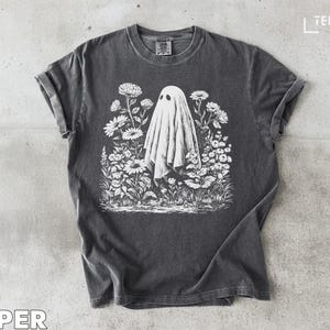 Puede incluir: Camiseta gris oscuro con una ilustración blanca de un fantasma en un campo de flores. La camiseta tiene cuello redondo y mangas cortas. La palabra "TEEZARO" está impresa en la esquina superior derecha.