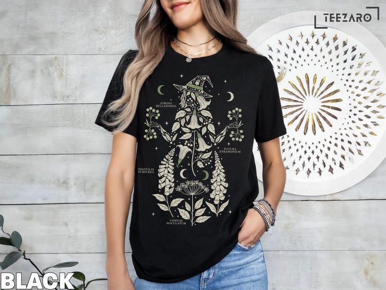 Puede incluir: Camiseta negra con un dise&ntilde;o de bruja bot&aacute;nica. El gr&aacute;fico incluye una bruja con sombrero, rodeada de ilustraciones de plantas y texto. El dise&ntilde;o incorpora lunas crecientes y estrellas, creando un tema m&iacute;stico.