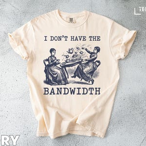 Puede incluir: Camiseta de color marfil con una ilustración vintage de dos mujeres en una mesa, con el texto "I DON'T HAVE THE BANDWIDTH" en negro. El gráfico representa una escena de fiesta de té con tazas y una tetera.