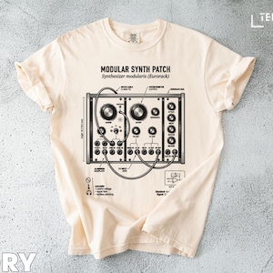 May include: Ivory-colored t-shirt featuring a detailed black and white illustration of a modular synthesizer patch. The design includes knobs, jacks, and text that reads "Modular Synth Patch" and "Synthesizer modularis (Eurorack)."