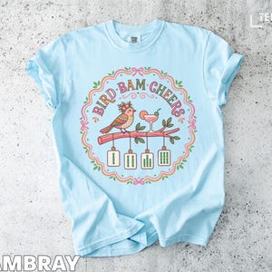 Light blue t-shirt with "Bird, Bam, Cheers" text and illustration of birds with cocktails.