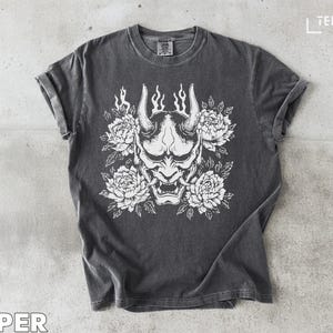 May include: Dark gray t-shirt featuring a white graphic of a horned demon mask surrounded by white peony flowers. The shirt has rolled-up sleeves and the word "PEPPER" is printed at the bottom.