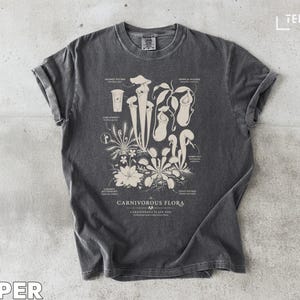 Puede incluir: Una camiseta gris oscuro con una ilustración botánica detallada de plantas carnívoras. El diseño incluye varias especies de plantas con etiquetas y el texto "CARNIVOROUS FLORA". La camiseta tiene un aspecto vintage.