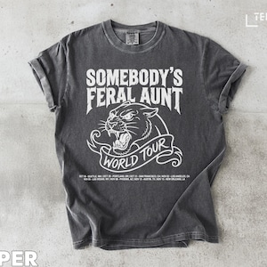 Peut inclure: T-shirt couleur poivre avec des manches retroussées. Le t-shirt affiche le texte "SOMEBODY'S FERAL AUNT" au-dessus d'un graphique de panthère rugissante et "WORLD TOUR" sur une bannière. Les dates de la tournée sont listées en dessous.