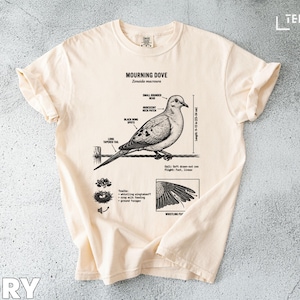 Op de afbeelding: Ivoor T-shirt met een gedetailleerde illustratie van een rouwduif, met anatomische labels. Het ontwerp toont de afmetingen van de vogel, het nest en vleugels. Het shirt heeft een ronde hals en korte mouwen.