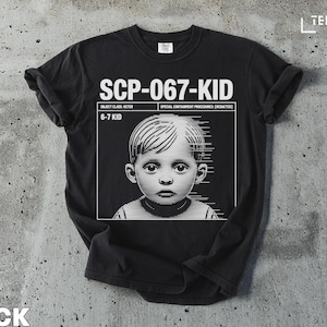 May include: Black t-shirt featuring the text "SCP-067-KID" above a grayscale image of a child. The shirt also includes the text "OBJECT CLASS: KETER" and "6-7 KID". The brand name "TEEZARO" is visible.