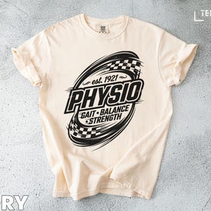 Puede incluir: Camiseta de color marfil con un diseño gráfico negro. El diseño presenta la palabra "Physio" dentro de un óvalo con un patrón de cuadros, junto con el texto "Est. 1921" y "Gait Balance Strength".
