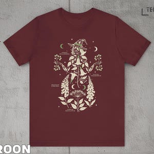 Puede incluir: Camiseta granate con una ilustraci&oacute;n bot&aacute;nica de una bruja, con elementos florales y celestiales. El dise&ntilde;o incluye etiquetas de texto y un sombrero de bruja. La camiseta est&aacute; hecha de un material suave.