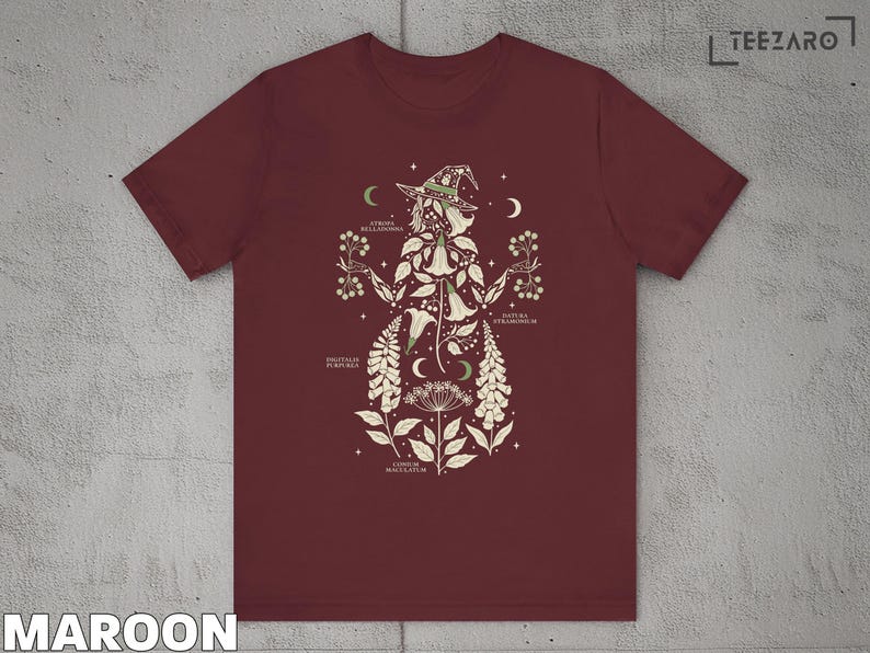 Puede incluir: Camiseta granate con una ilustraci&oacute;n bot&aacute;nica de una bruja, con elementos florales y celestiales. El dise&ntilde;o incluye etiquetas de texto y un sombrero de bruja. La camiseta est&aacute; hecha de un material suave.