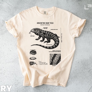May include: Ivory t-shirt with a black and white illustration of an Argentine B&W Tegu lizard. The design includes anatomical details and the text "ARGENTINE B&W TEGU". The shirt is made of a soft material.
