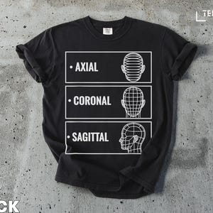 Puede incluir: Camiseta negra con un diseño gráfico blanco que presenta tres secciones etiquetadas: Axial, Coronal y Sagital, cada una con una ilustración de cabeza. La palabra "BLACK" está en la parte inferior.
