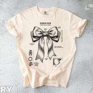 May include: Ivory-colored t-shirt featuring a detailed black and white illustration of a ribbon bow with labeled parts. The text "RIBBON BOW" is at the top, with additional diagrams and text below. The word "IVORY" is at the bottom.