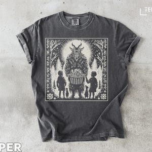 May include: Dark gray t-shirt featuring a black and white graphic of a horned creature holding a basket with two children and two dogs. The design is framed with a border of text and snowflakes.