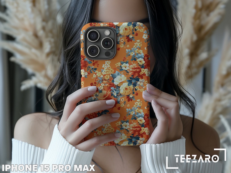 May include: A floral phone case for an iPhone 15 Pro Max. The case has a bright orange background with a colorful floral pattern. The case is being held by a person, but only the hands are visible.