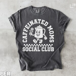 Puede incluir: Camiseta de color pimienta con un gráfico blanco. El gráfico presenta una tetera de dibujos animados sosteniendo una flor, con las palabras "CAFFEINATED MOMS SOCIAL CLUB" en semicírculo arriba y abajo de la imagen. La camiseta tiene mangas cortas y cuello redondo.