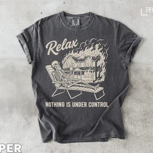 May include: Dark gray t-shirt with a graphic of a skeleton relaxing in a beach chair while a house burns in the background. The text "Relax" and "Nothing is Under Control" are printed on the shirt.